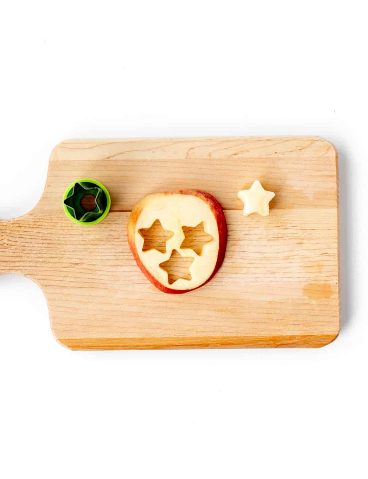 A star shaped cookie cutter on a wooden cutting board, with stars cut out of an apple.