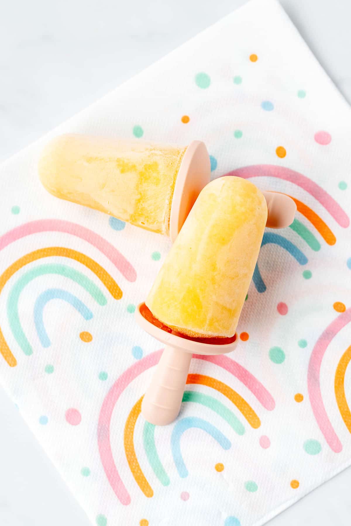 Two mango popsicles lying flat on a rainbow tablecloth.