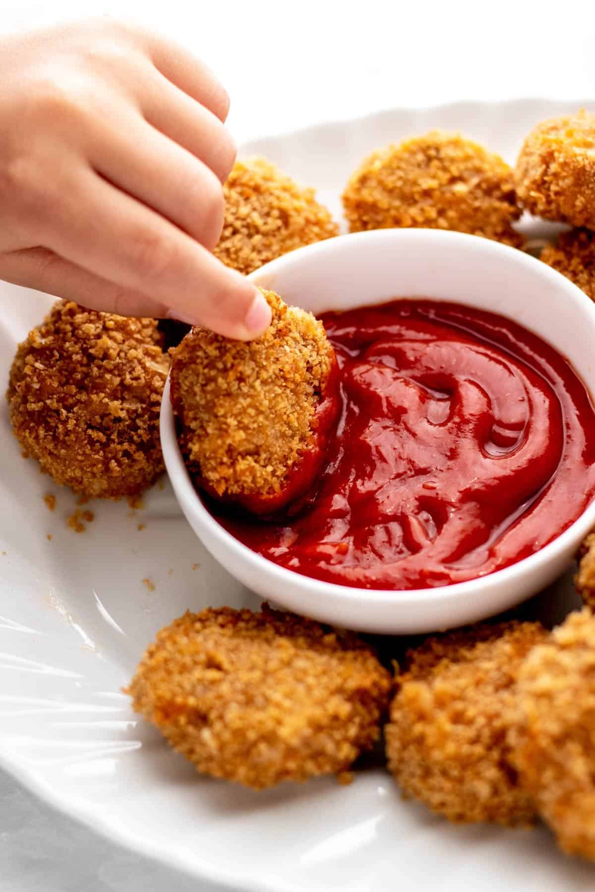 Air fryer chicken nuggets on a plate, with someone dipping one into a bowl of ketchup.