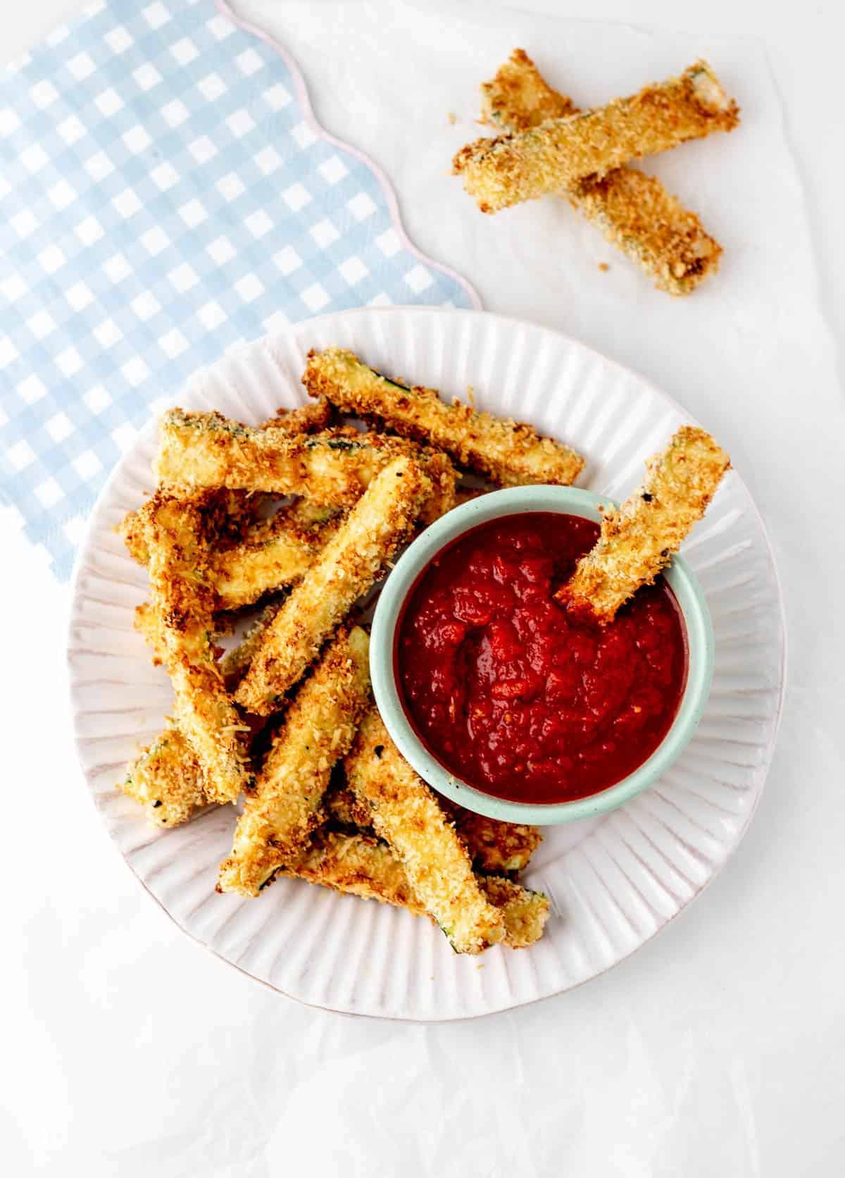 Air fryer zucchini fries on a white plate, with one fry being dipped into a bowl of sauce.