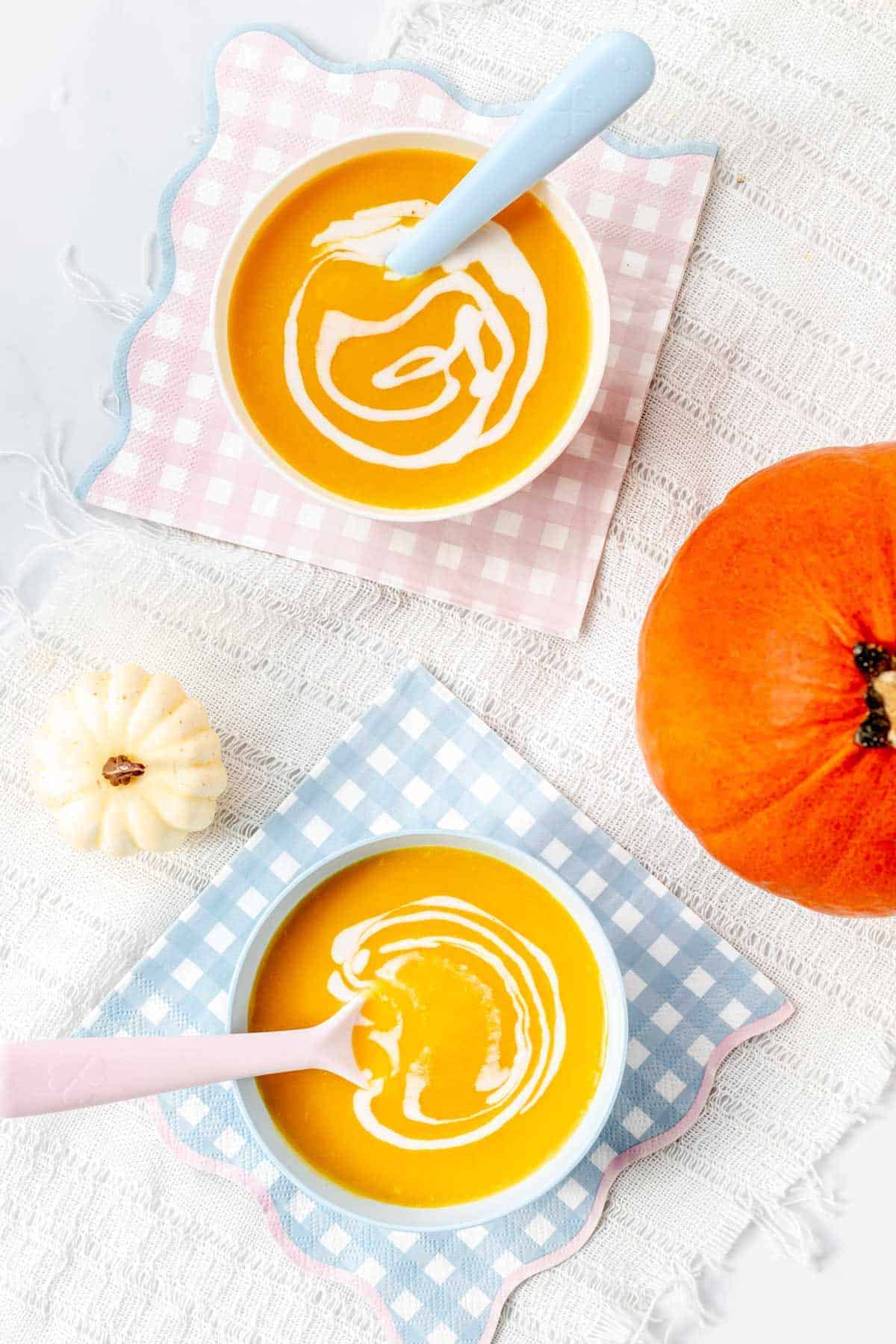 Two small white bowls of pumpkin soup on plaid napkins next to a pumpkin.