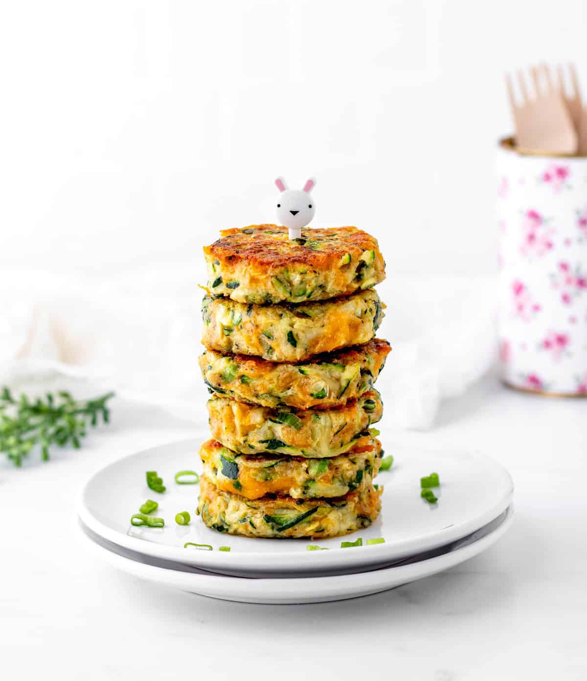 Cheesy zucchini fritters stacked on top of one another on a white plate with a bunny toothpick on top.