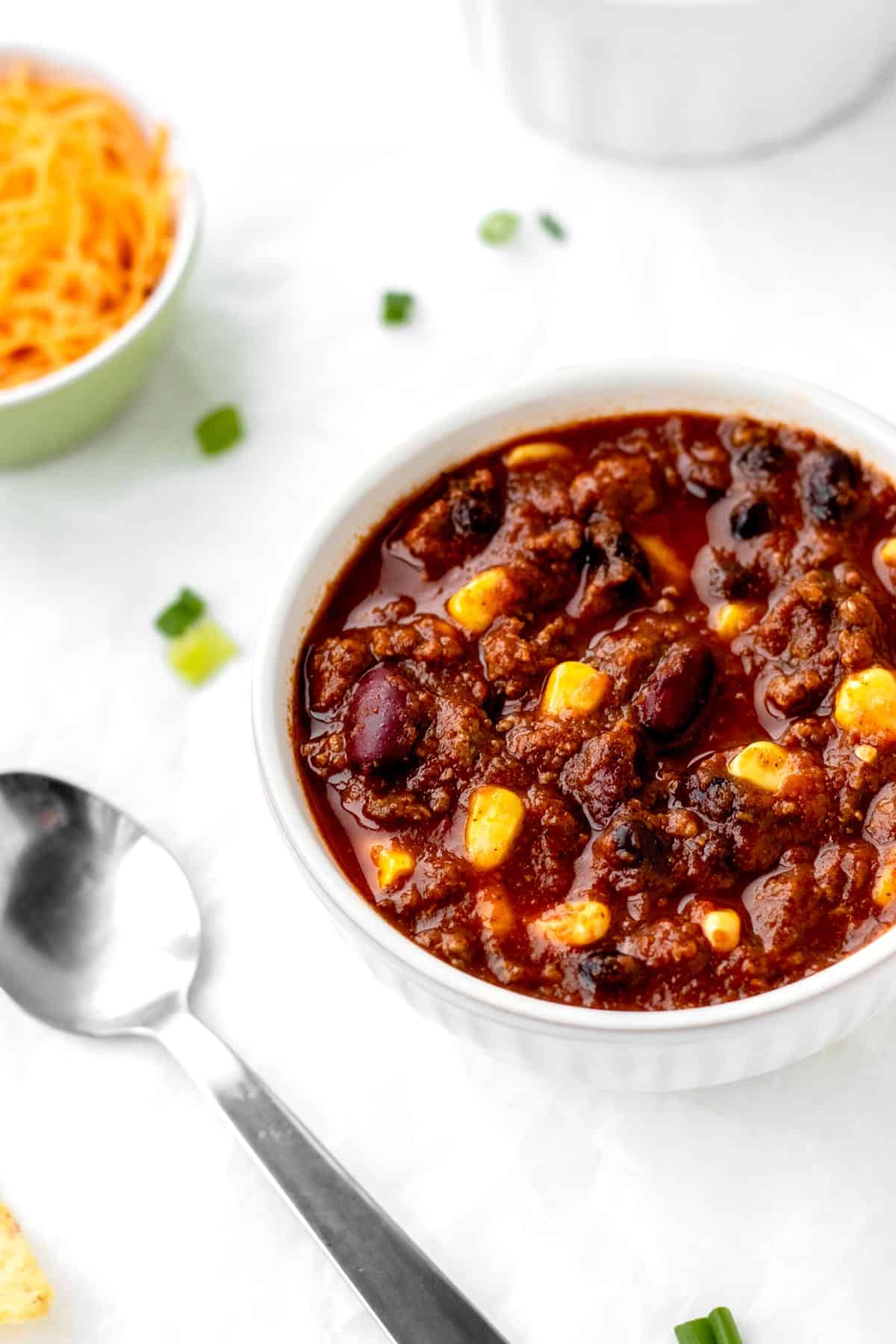 A small bowl of chili con carne on a table, next to a spoon and a bowl of shredded cheese.