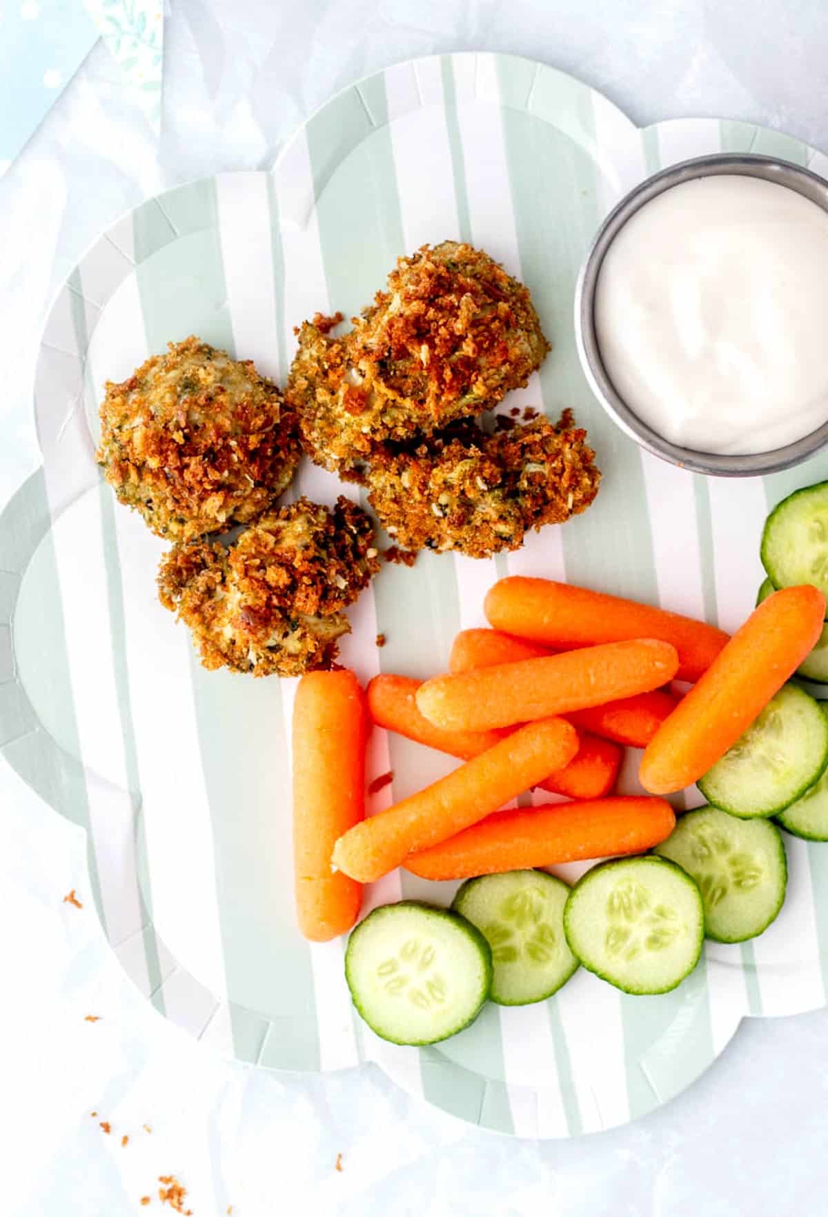 Pesto chicken bites with baby carrots and cucumber slices on a blue striped plate.