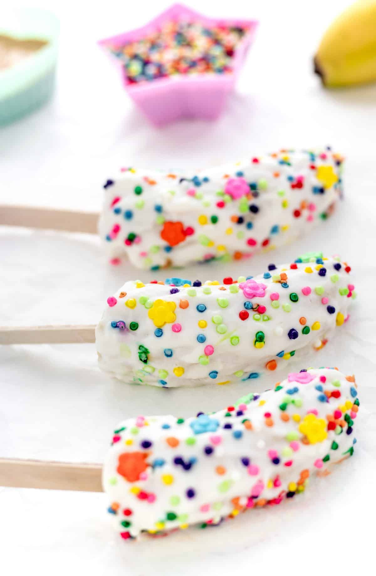3 frozen banana pops with yogurt, decorated in colorful sprinkles, on a baking sheet.
