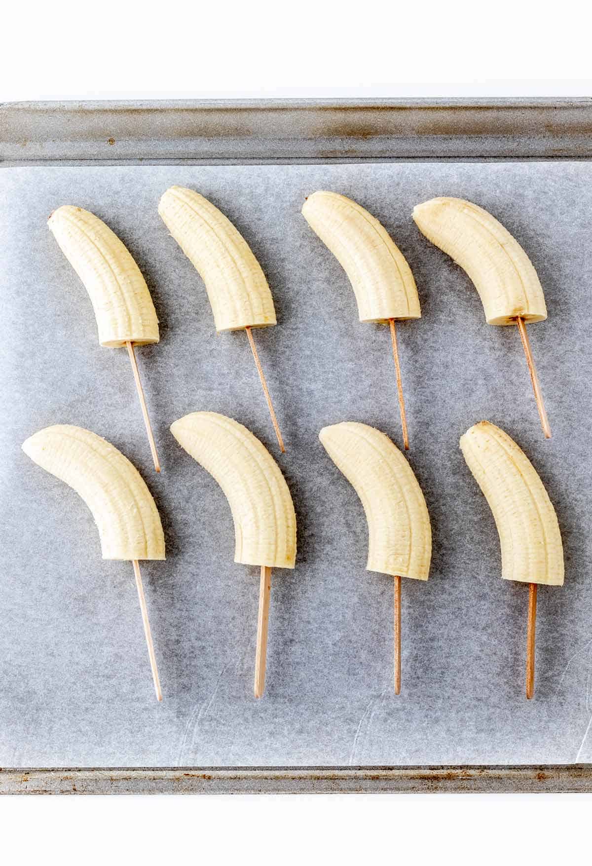 8 bananas with popsicle sticks on a parchment paper lined baking sheet.
