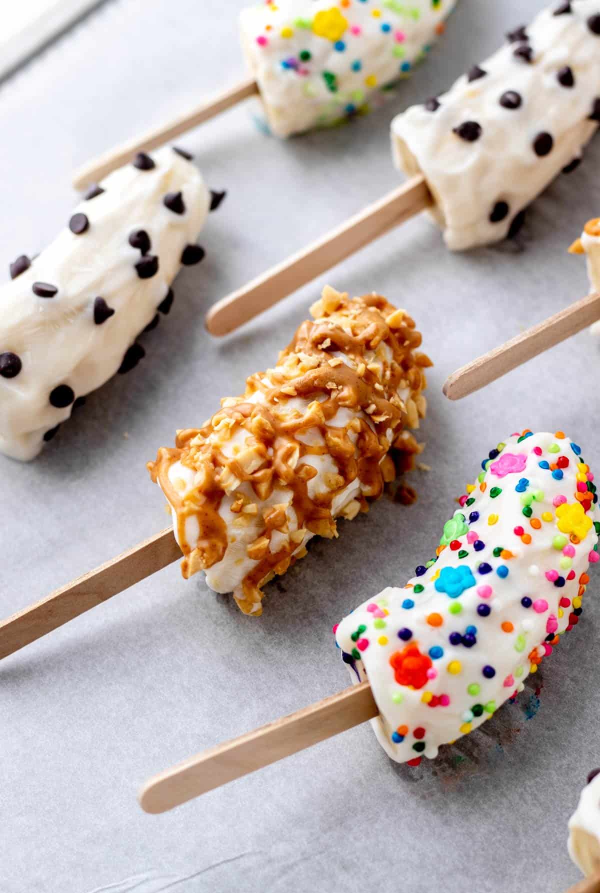 A close-up of the frozen banana pops with yogurt covered in various toppings on a parchment paper lined baking sheet.