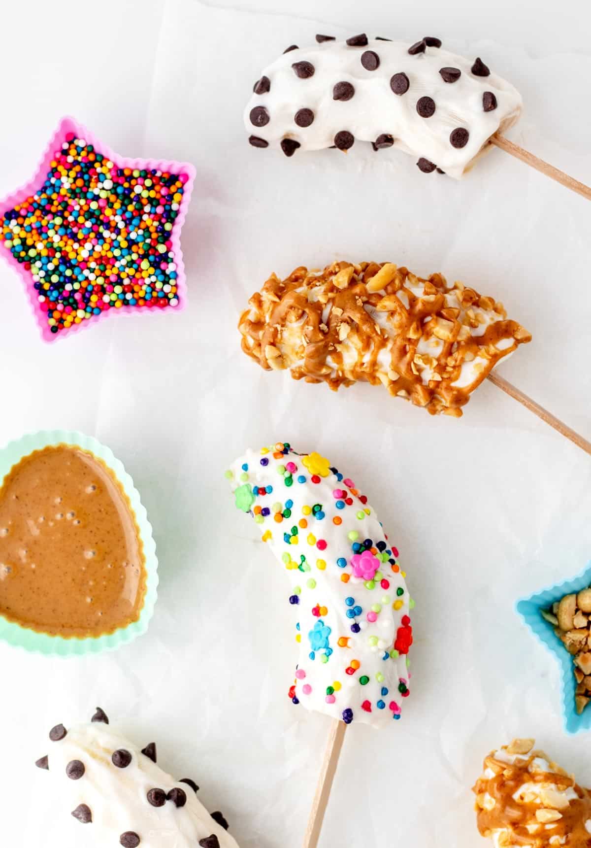Decorated frozen banana yogurt pops on a baking sheet surrounded by a bowl of sprinkles and a bowl of peanut butter for decorating.
