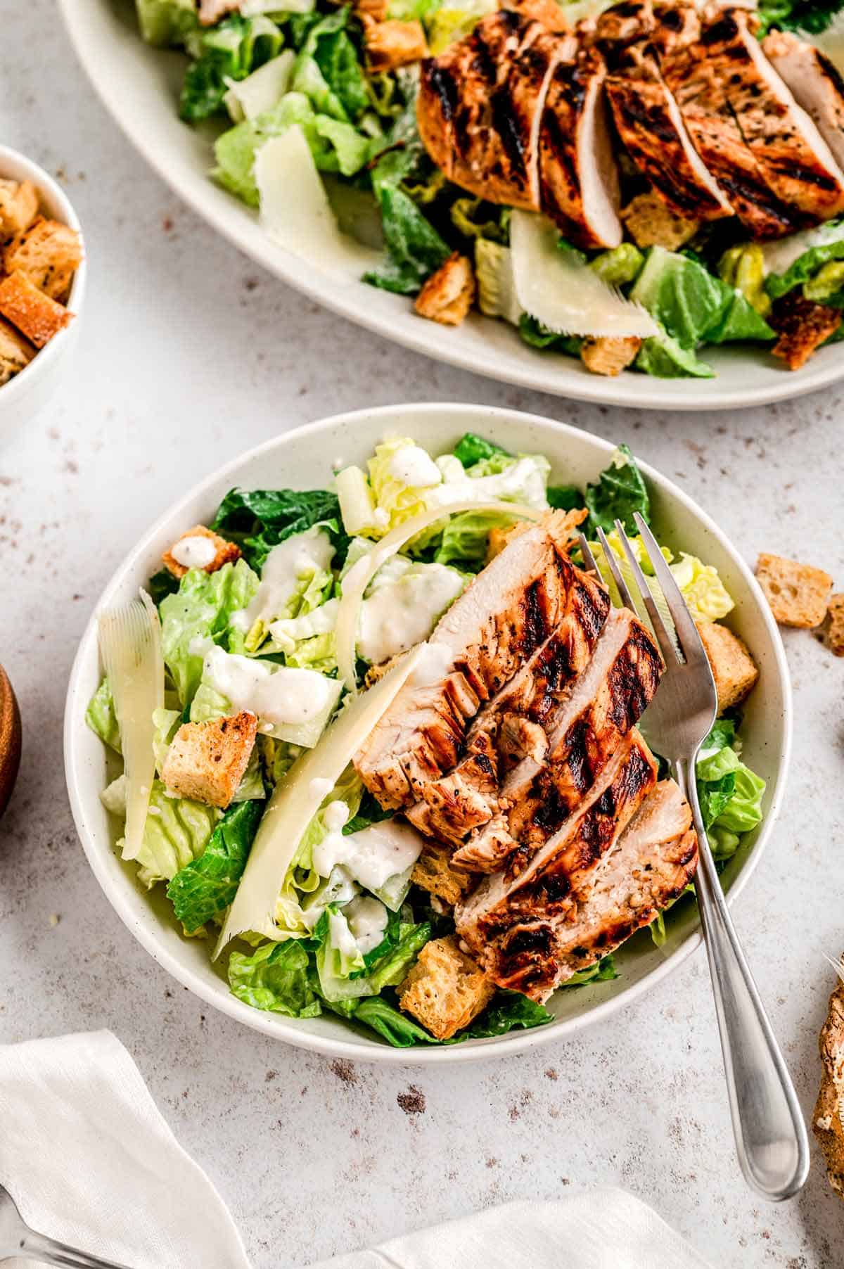 A bowl of chicken Caesar salad on a table next to a large salad bowl.