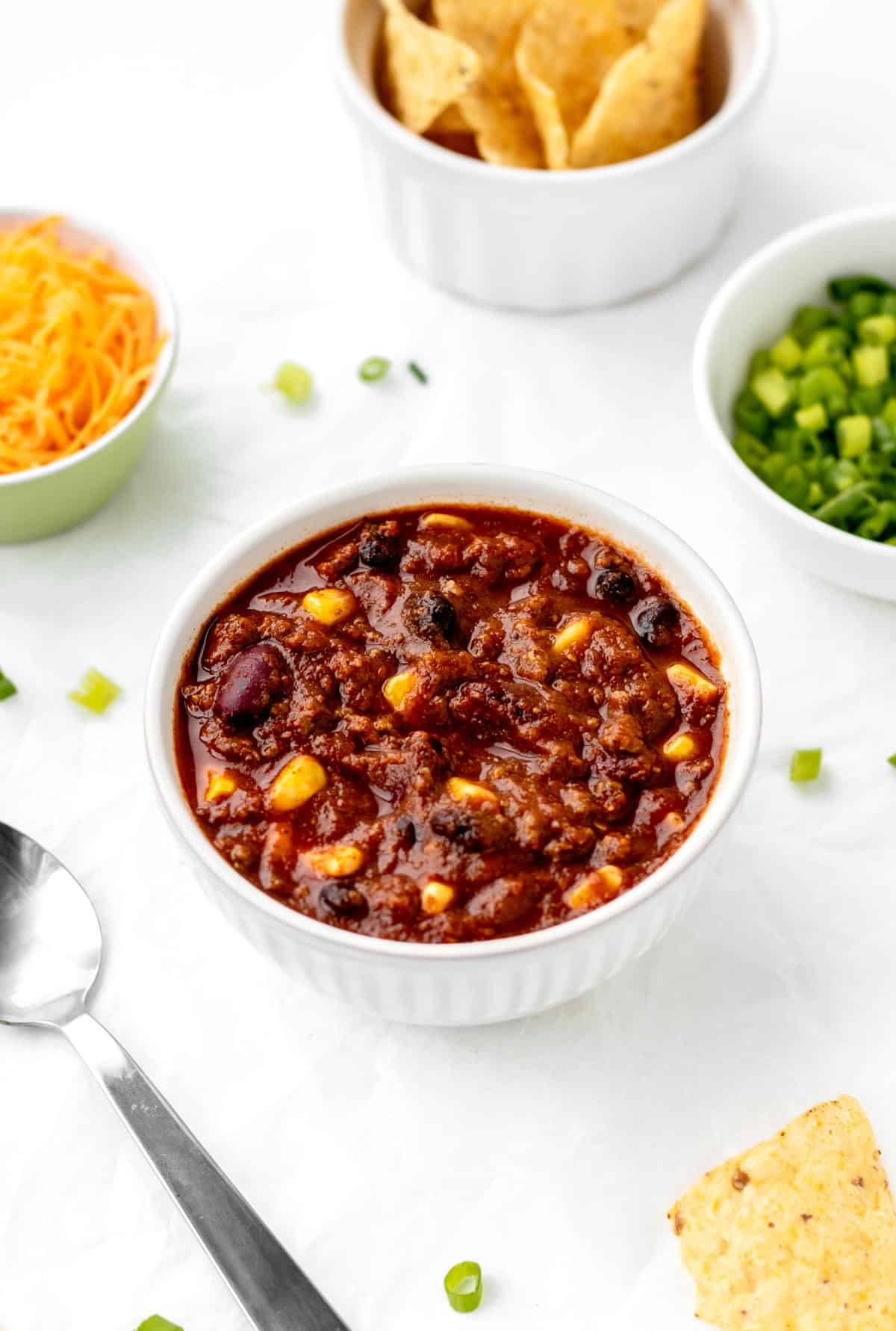 A bowl of kid friendly chili con carne on a table next to a spoon, a bowl of green onions and shredded cheese.