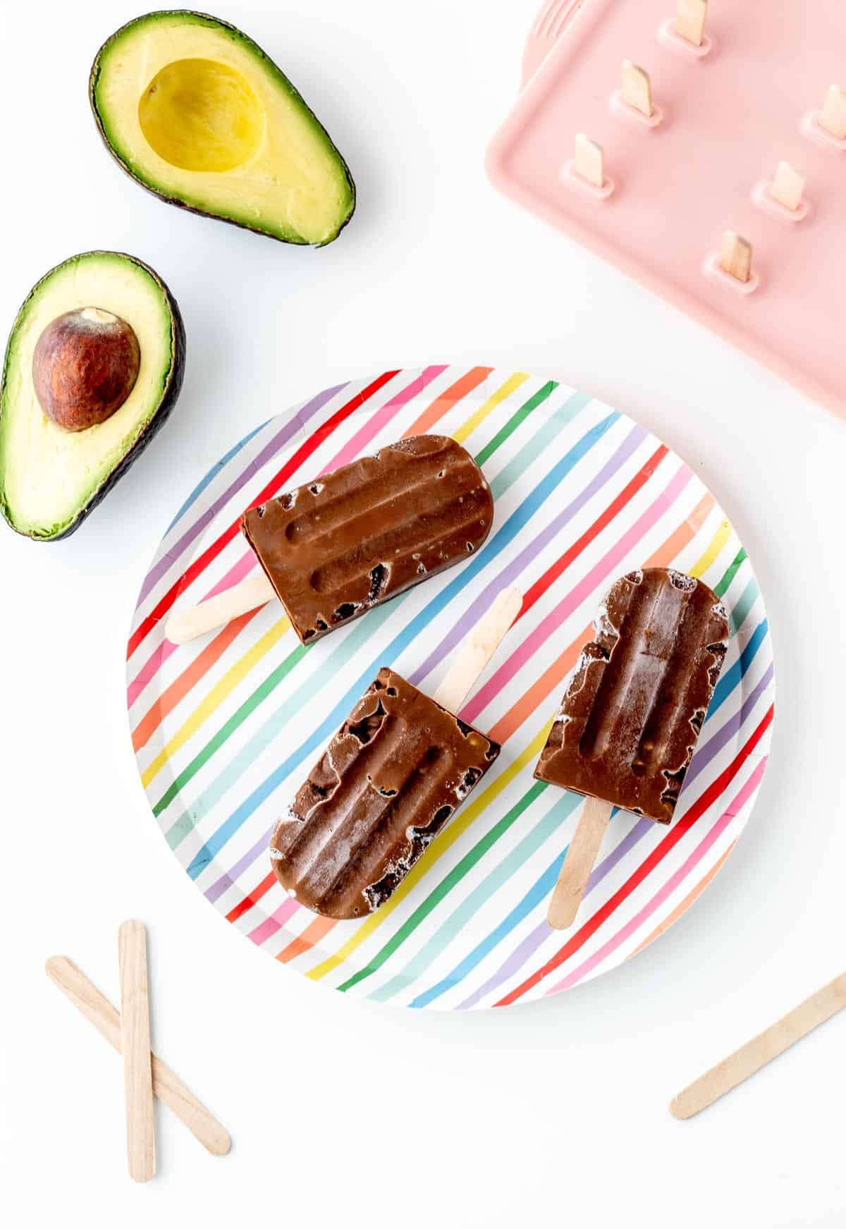 Three chocolate avocado popsicles on a colorful striped plate on a counter.