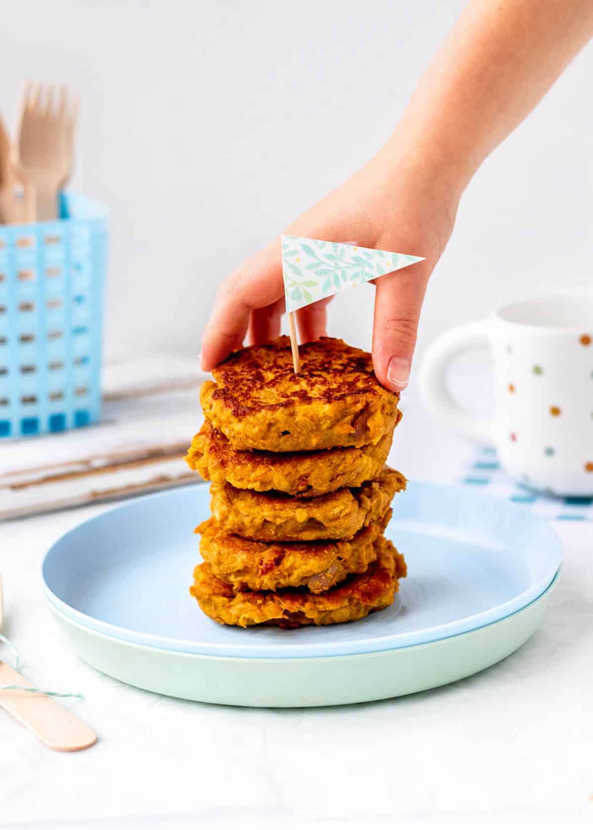 A child's hand reaching for a fritter on a stack of mashed sweet potato fritters.