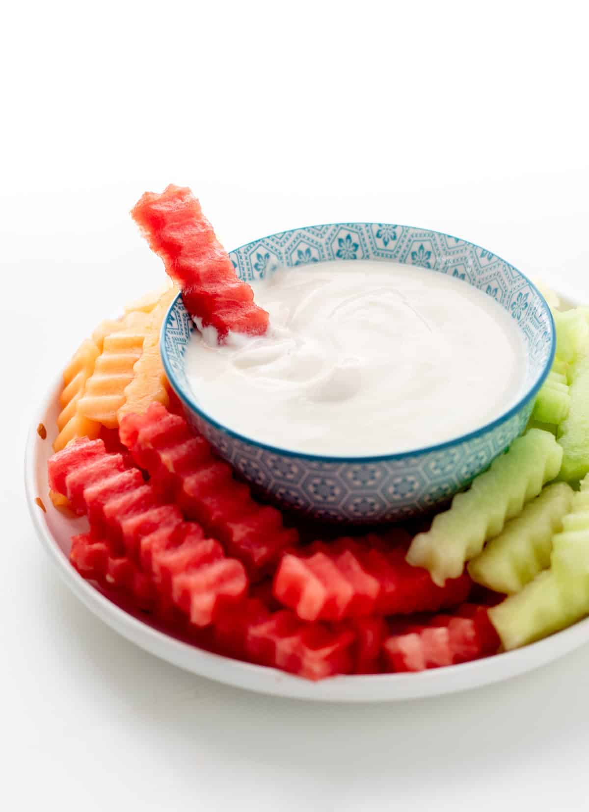 A plate of fruit fries with someone dipping a watermelon fry into the yogurt dip in the middle of the plate.