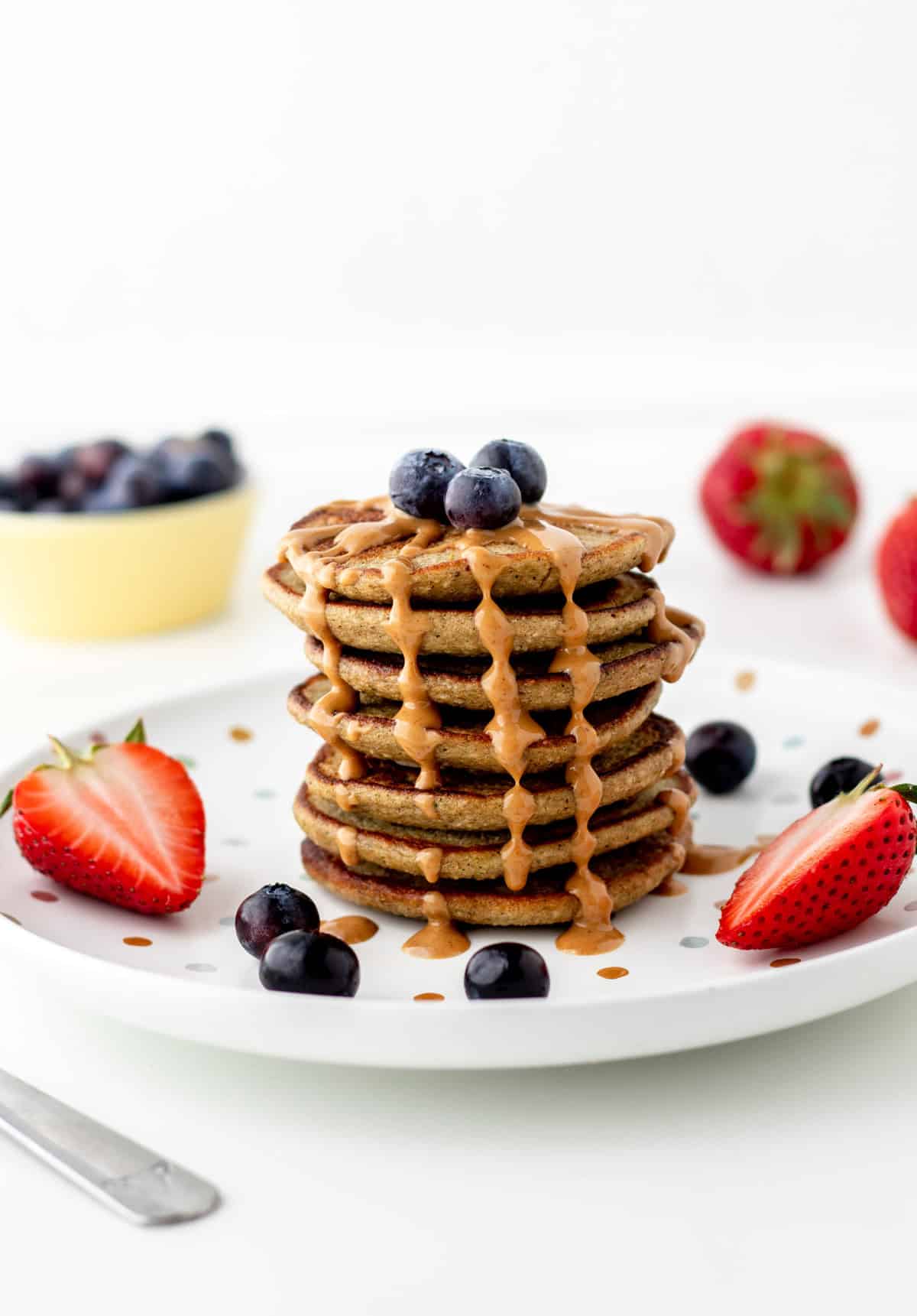 A stack of banana zucchini pancakes on a plate, topped with berries and a drizzle of peanut butter down the sides.