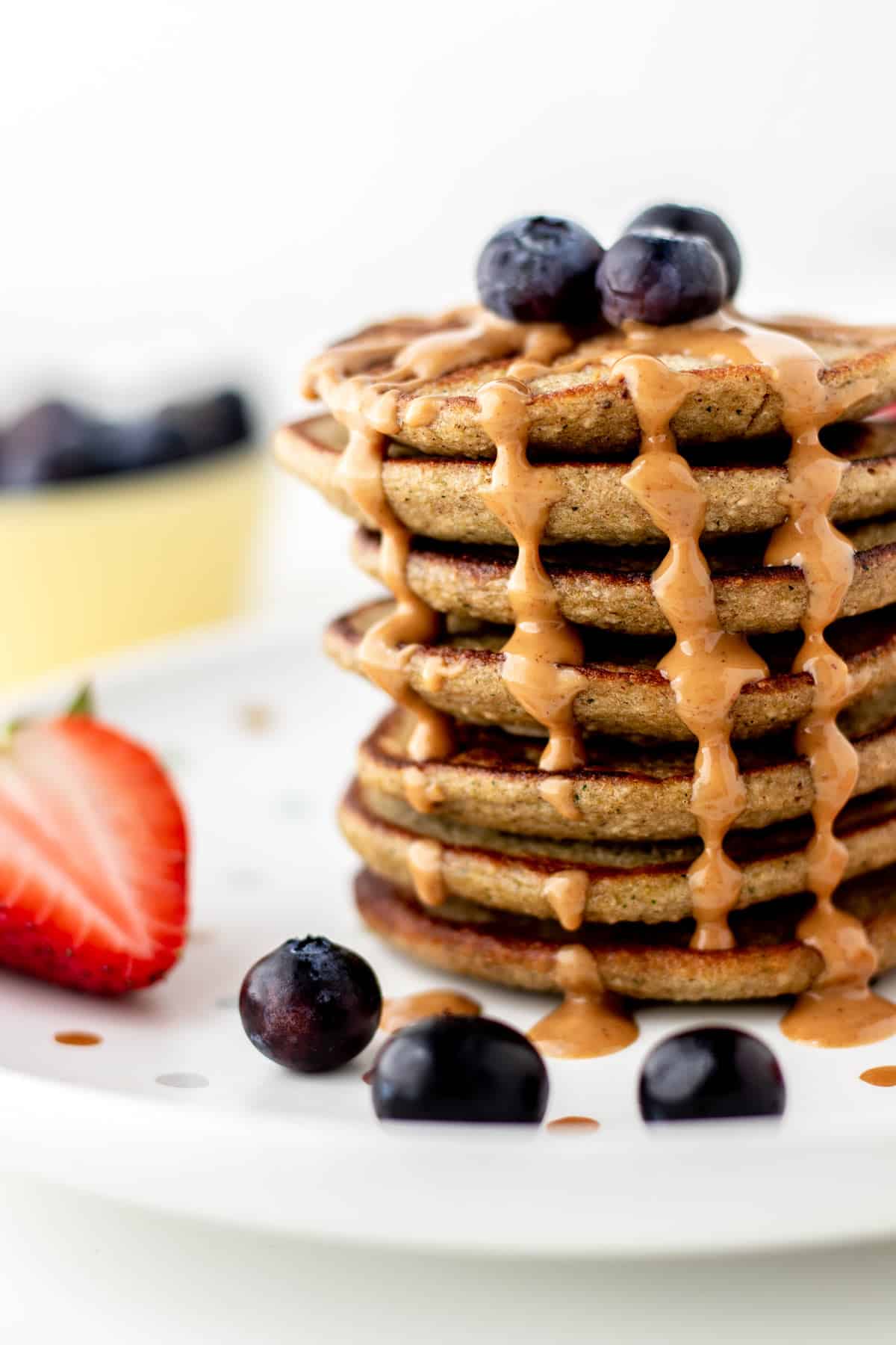 A large stack of banana zucchini pancakes with peanut butter drizzling over the top with berries on a plate.