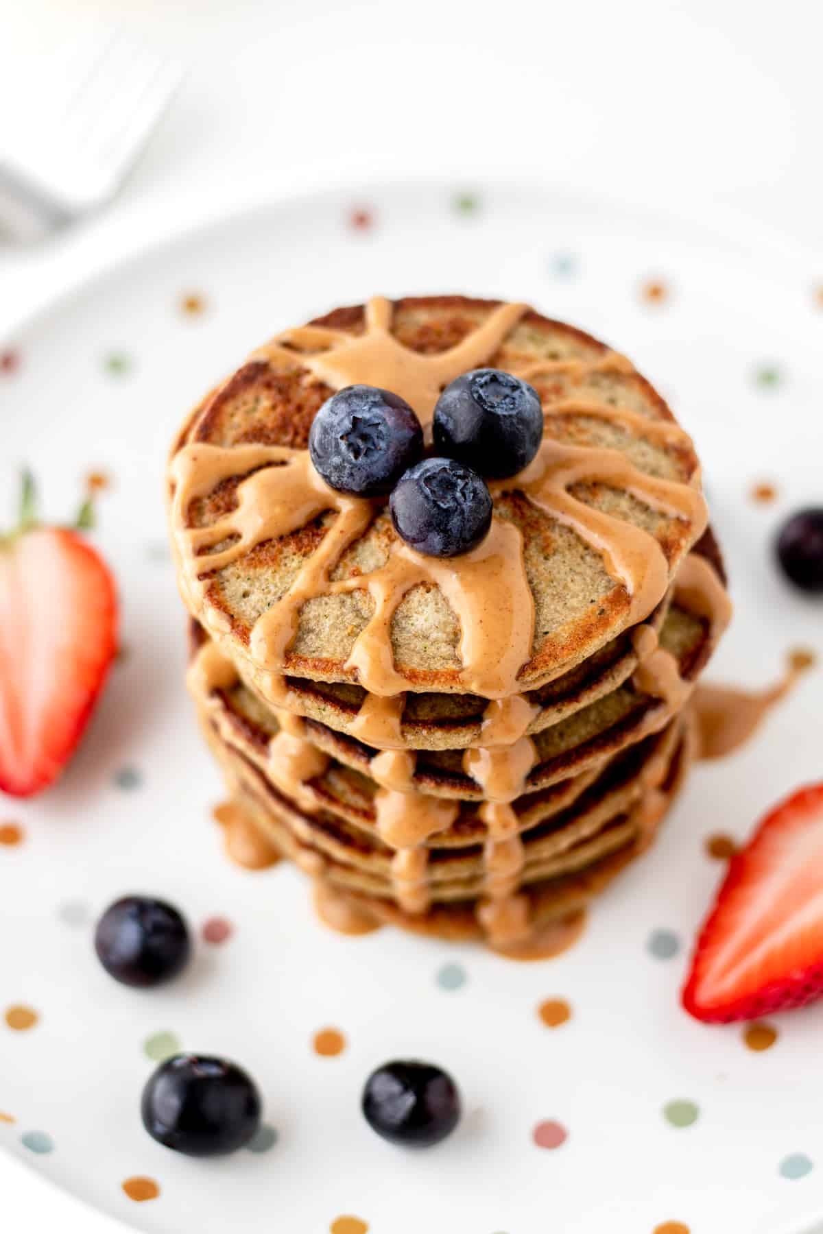 Banana zucchini pancakes stacked on top of one another on a polka dot plate and drizzled with peanut butter.