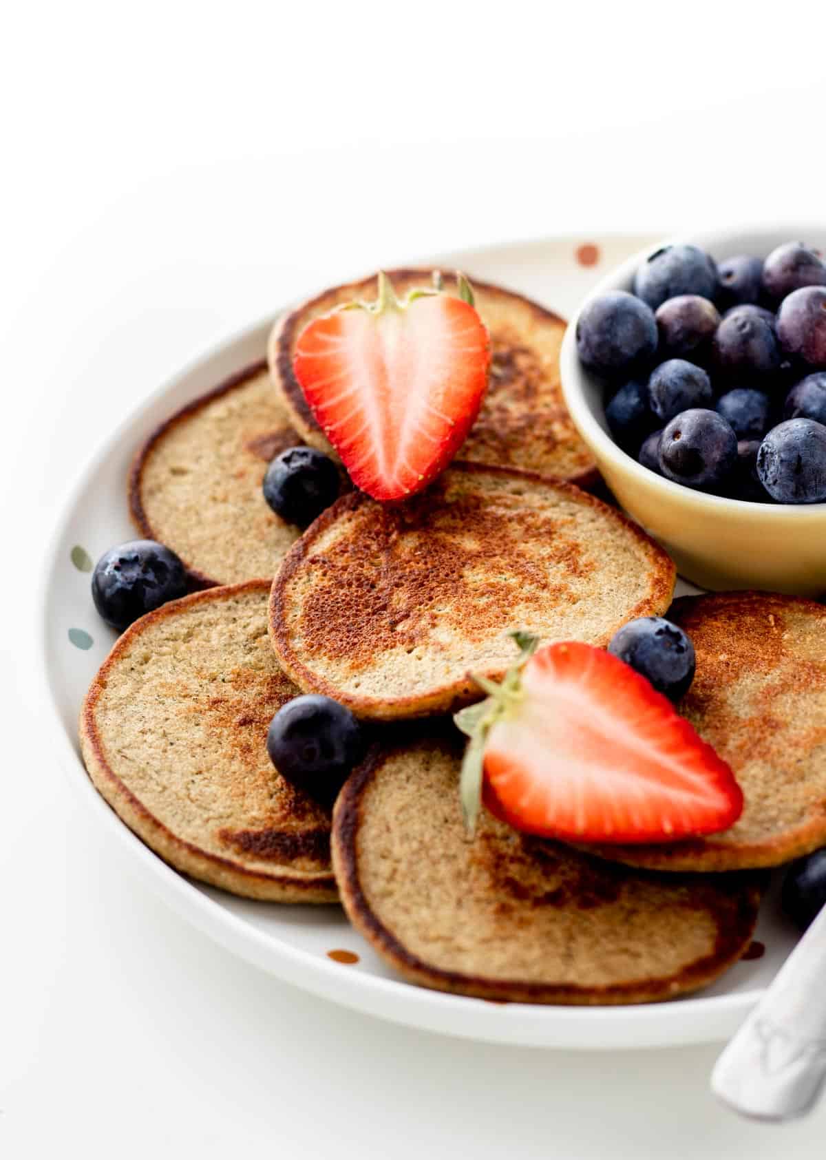 Banana zucchini pancakes on a plate next to a bowl of blueberries and topped with sliced strawberries.