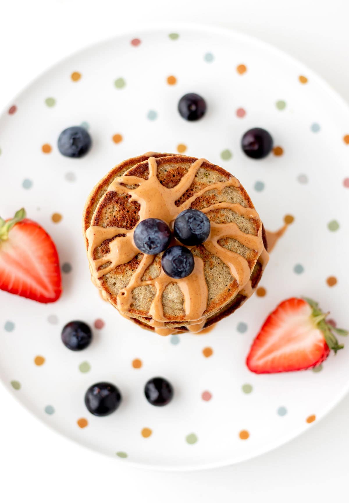 A birds-eye view of banana zucchini pancakes stacked on top of one another and drizzled with peanut butter on a polka dot plate.