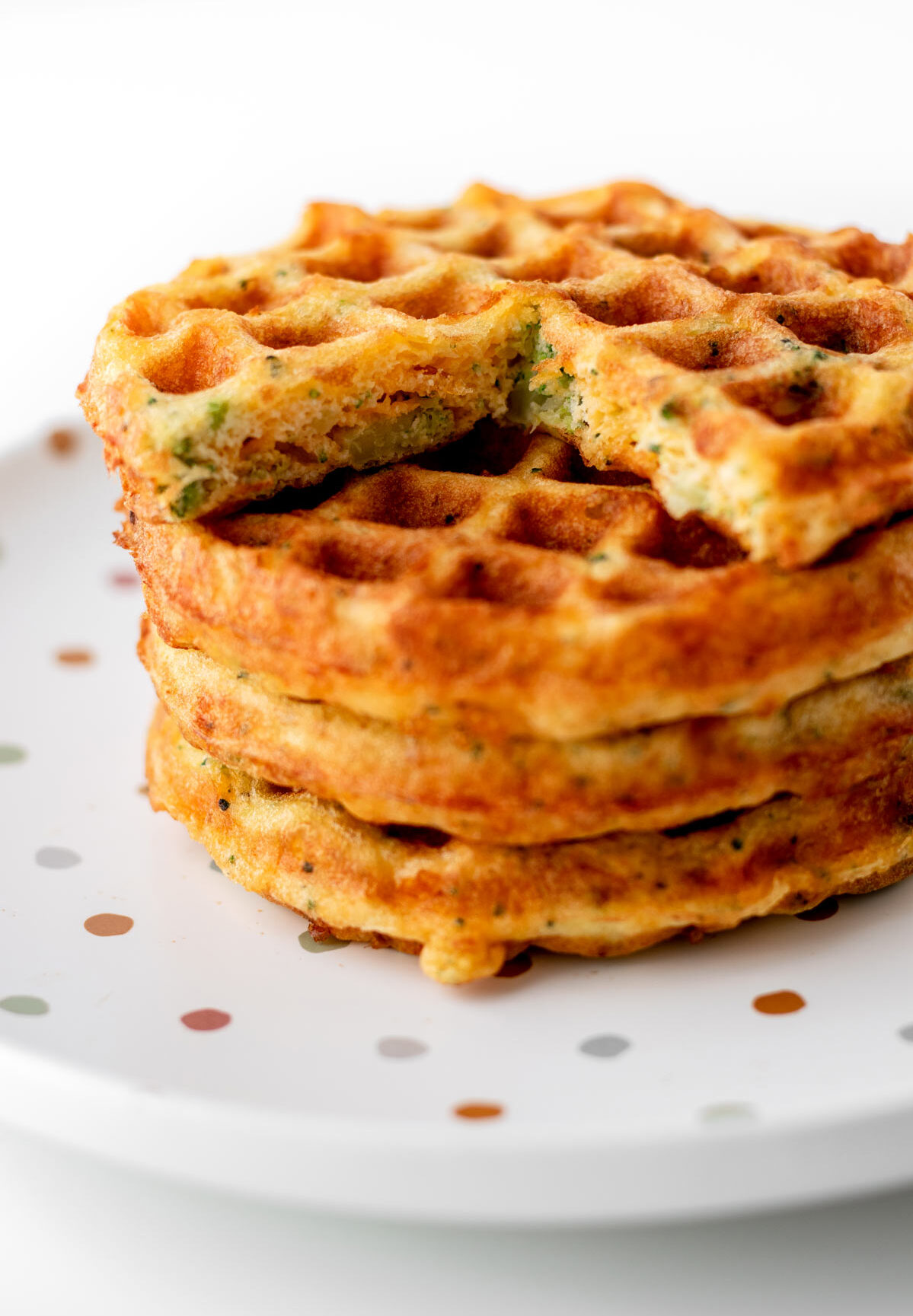 A stack of broccoli cheese waffles with the top waffle with a wedge cut out of it on a polka dot plate.