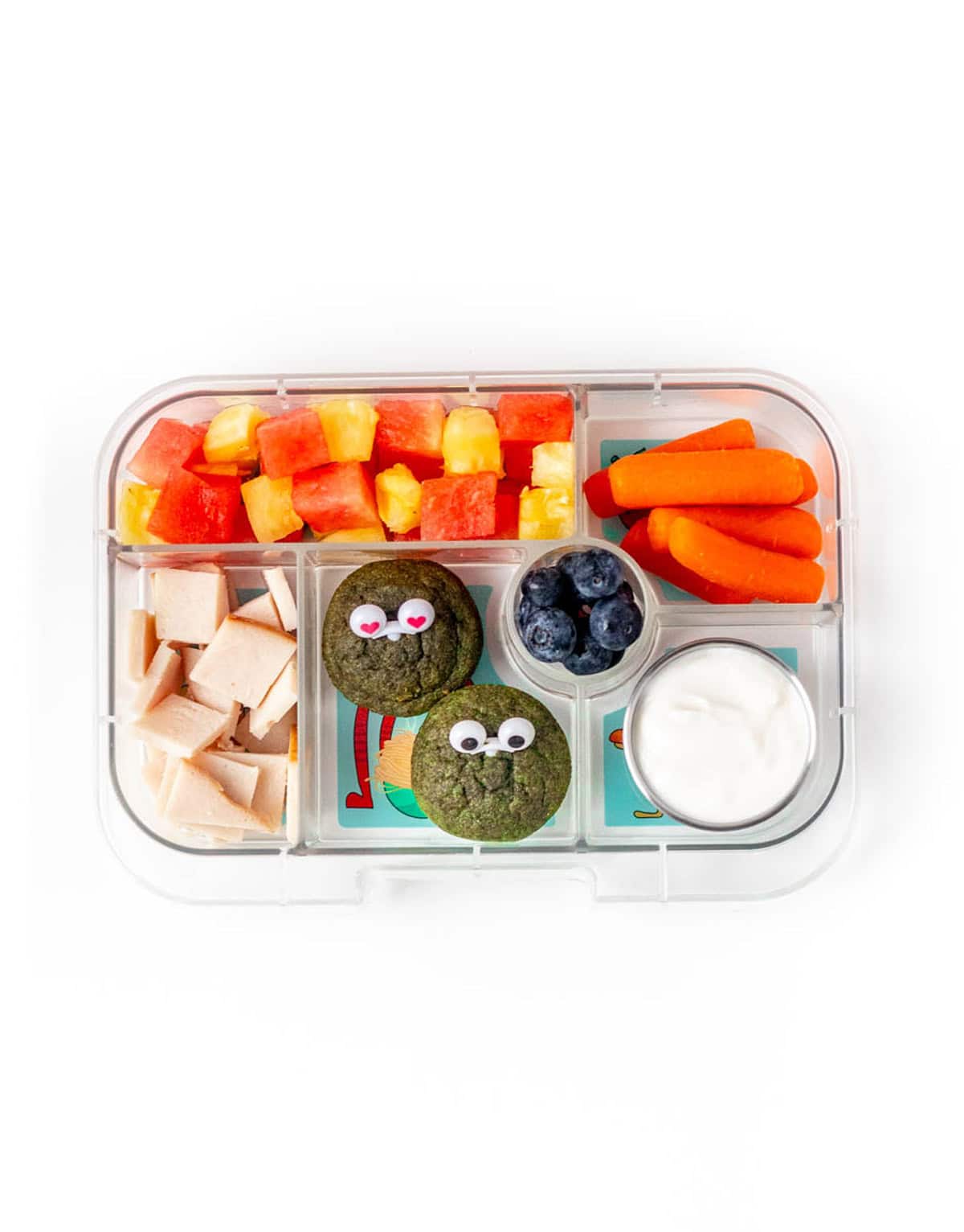 A lunchbox veggie idea featuring cubed melon, carrot sticks, blueberries, dip, sliced turkey and two green monster muffins with googly eyes on them.