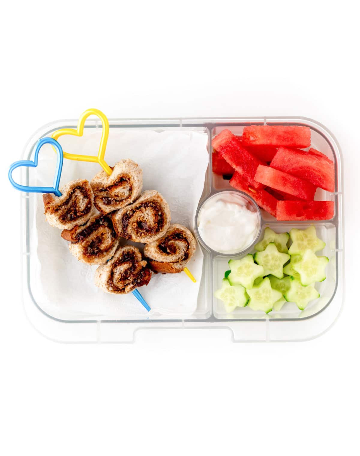 Mini cinnamon rolls on a heart skewer with watermelon sticks, cucumber stars and dip in a lunchbox.