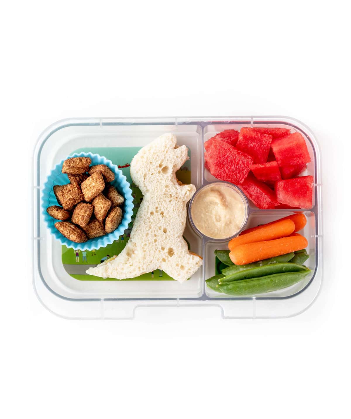 A lunchbox featuring a dinosaur sandwich, fruit and veggies with dip, and some cereal.