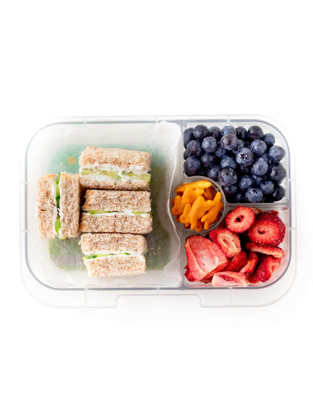 A lunchbox with mini cucumber sandwiches, fresh fruit and crackers.