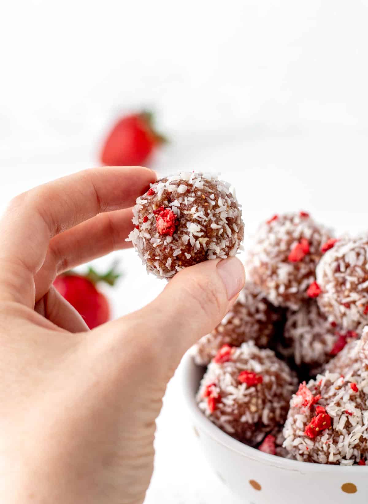 Someone holding up a strawberry bliss ball with a bowl of strawberry bliss balls in the background.