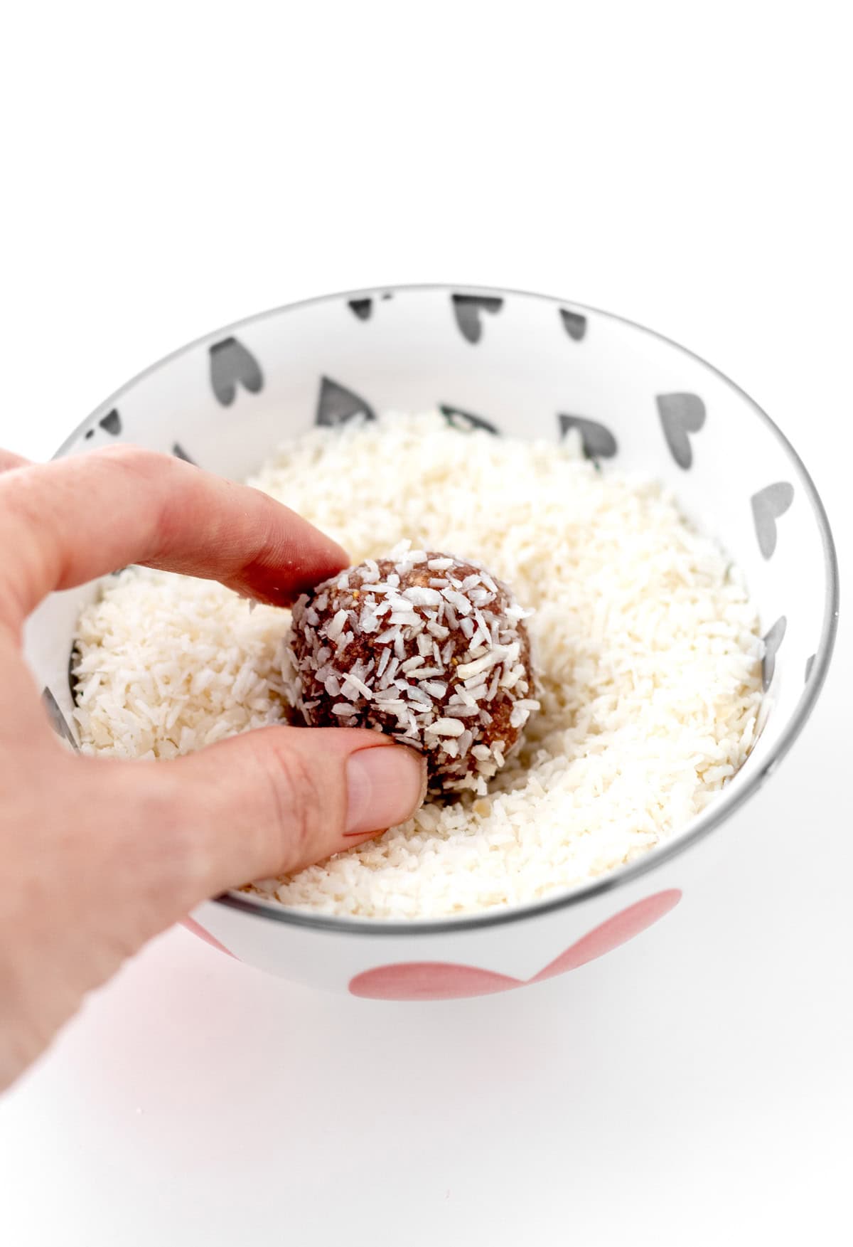 Someone holding a strawberry bliss ball, dipping it into a bowl of coconut flakes.