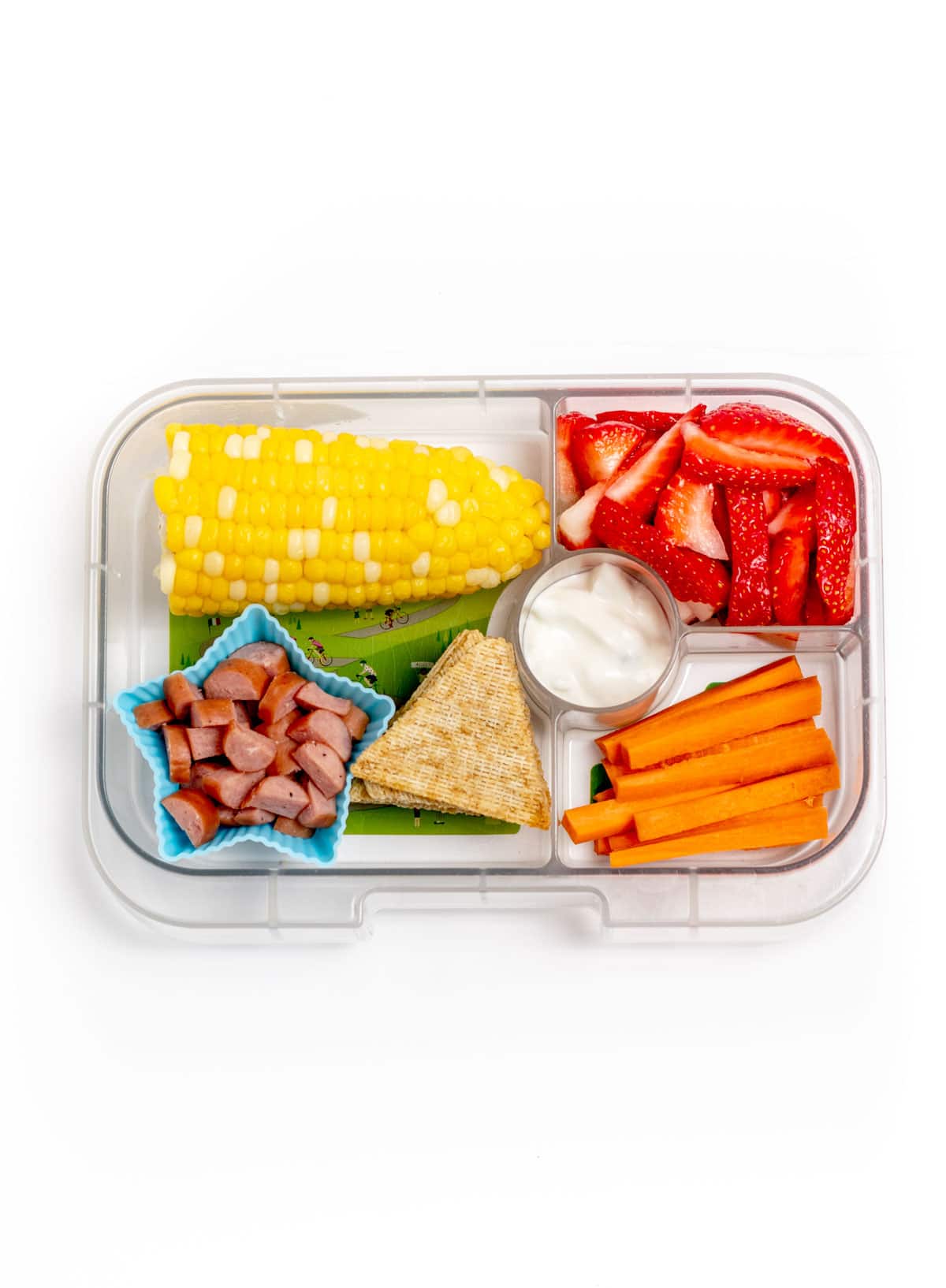 A lunchbox featuring a small corn on the cob, carrot sticks, bell peppers, pita, and dip.