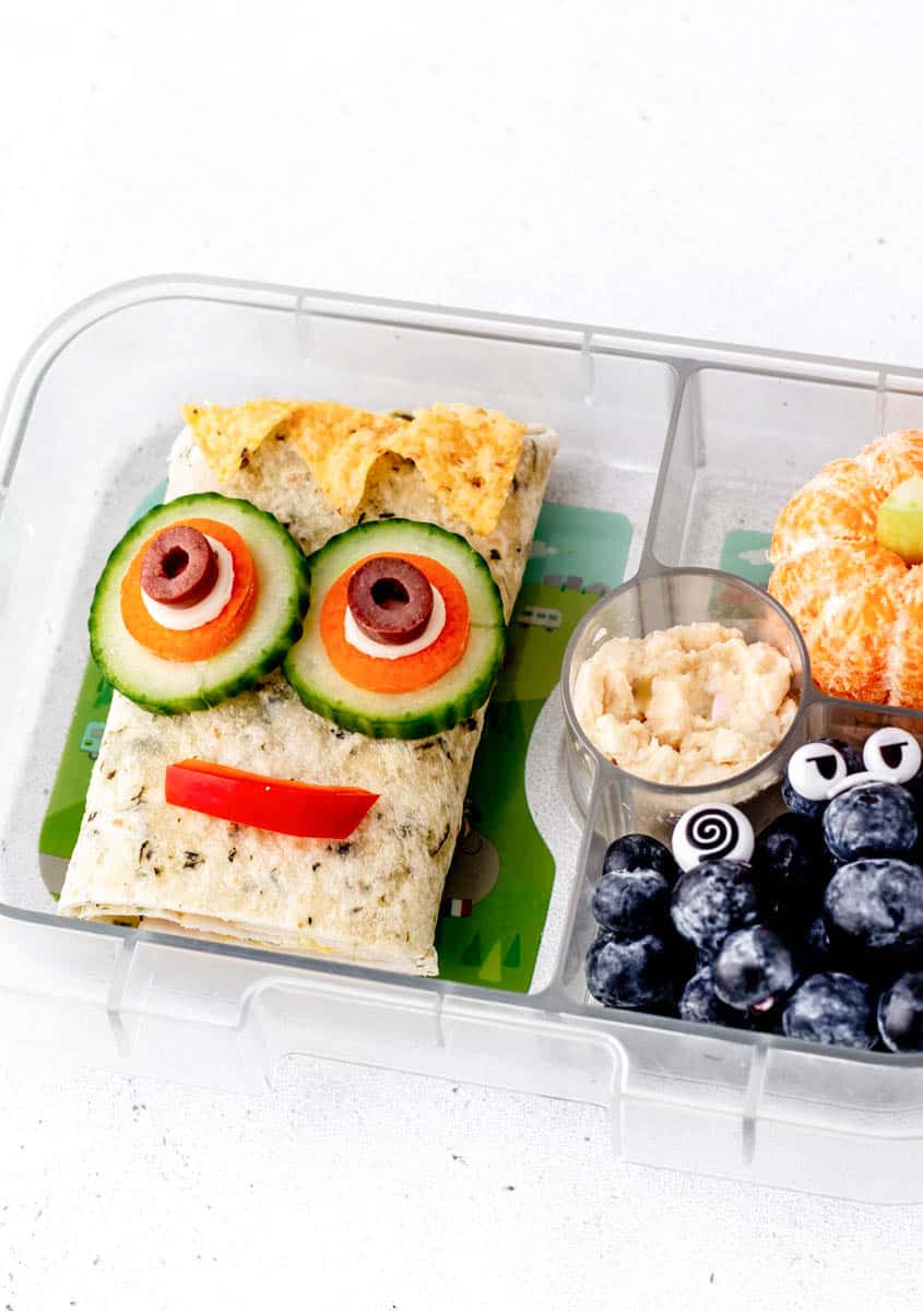 A pita sandwich in a lunch box with cucumber and olives that look like eyes.