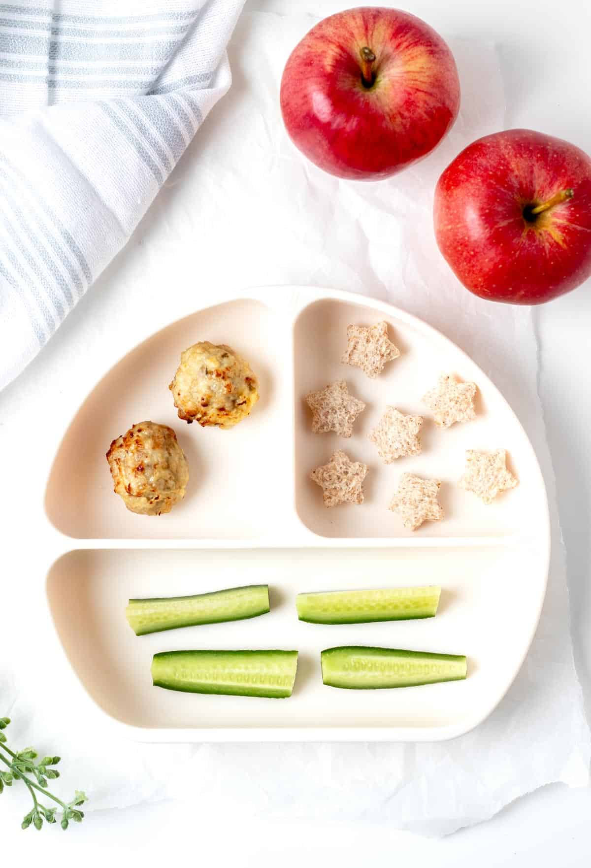 A divided plate with two chicken and apple meatballs, sliced cucumber, and bread cut out into the shape of stars.