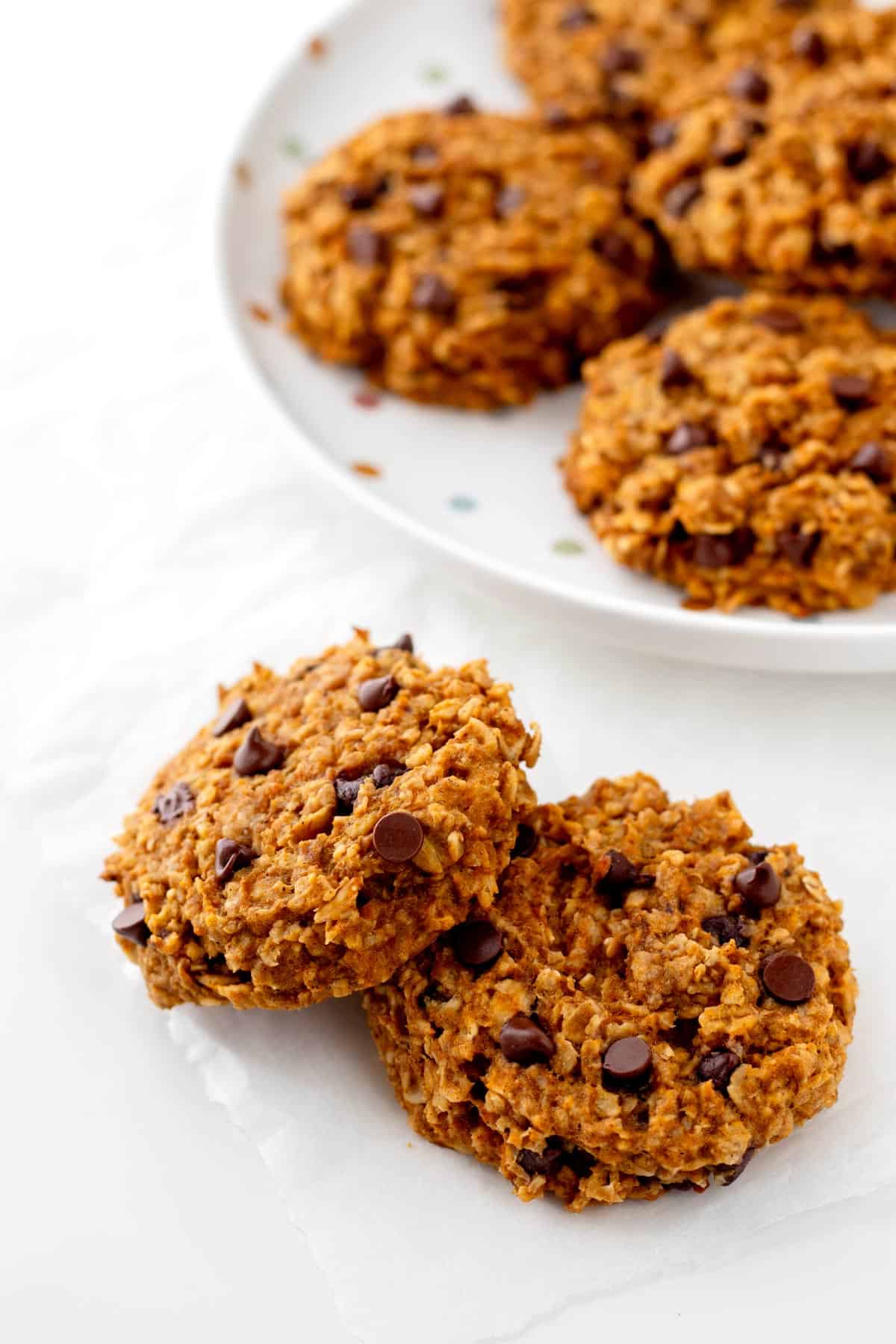 Two pumpkin banana cookies on a counter with a plate of cookies in the background.