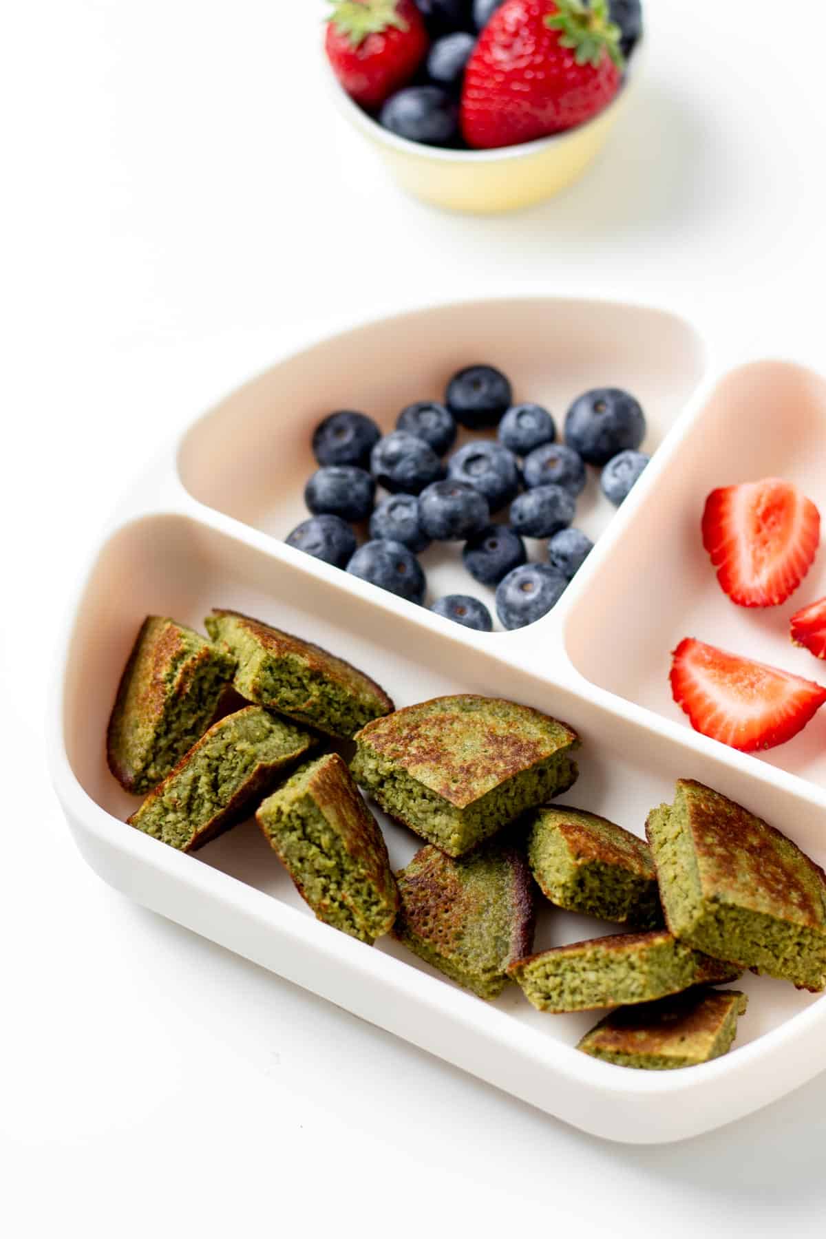 Cut up hulk pancakes on a divided plate with blueberries and sliced strawberries.