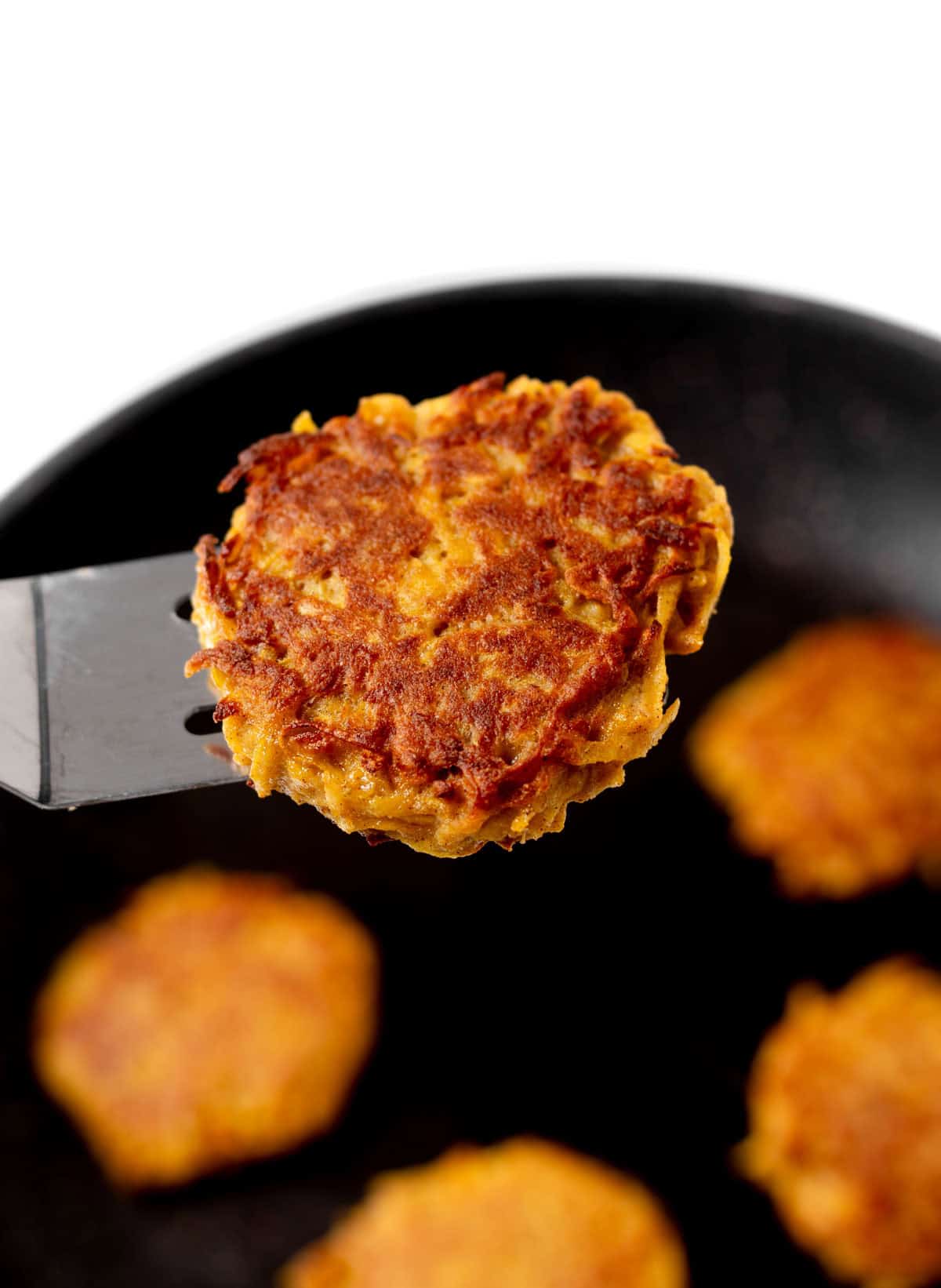 A close-up of a butternut squash cake on a spatula with a frying pan in the background.